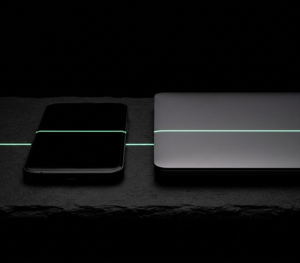 A dark smartphone and a closed laptop on black stone with a line of green light across both